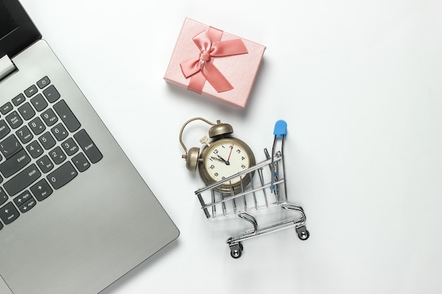 Computer portatile, sveglia retrò, carrello della spesa, scatole regalo con fiocco su sfondo bianco. 11:55. anno nuovo, concetto di natale. shopping in linea di vacanze. vista dall'alto