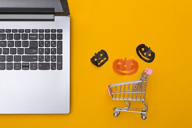 Laptop and shopping trolley with paper cut halloween pumpkins on a yellow background. Halloween theme. Top view