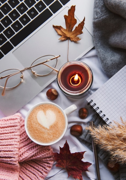 Laptop, sweater, yellow autumn leaves, candle and coffee on white bed. Work at home concept. Autumn, fall, winter composition. Flat lay, top view.