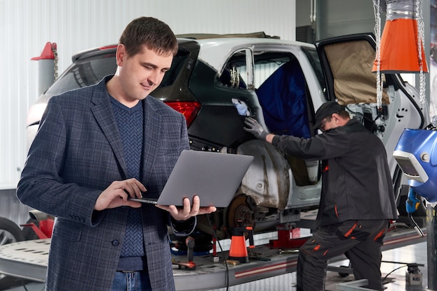 Laptop van de managerholding terwijl status dichtbij auto in autoreparatiewerkplaats met mechanische herstellende auto op achtergrond