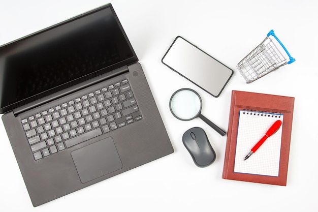 Laptop vergrootglas computermuis smartphone pen notitieblok en boodschappenmand op een witte achtergrond Artikelen voor het bedrijfsleven en het online kopen van goederen