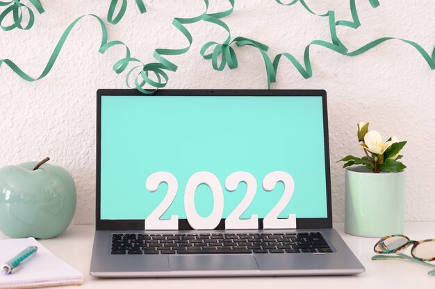 Laptop on white background with festive decoration, with number 2022 in wooden figures. Work, remote work, happy new year on the desk