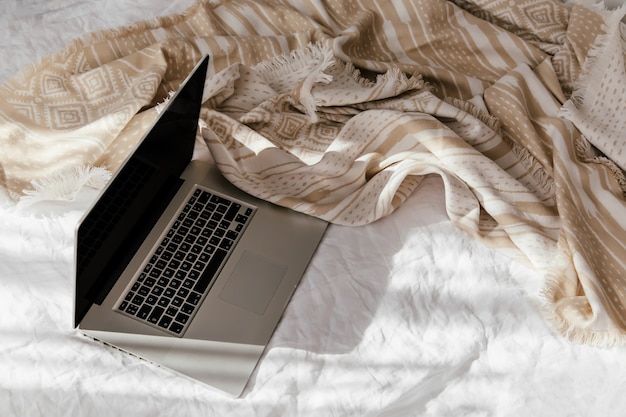 Laptop on white bed with a beige plaid. Work at home concept. Morning light