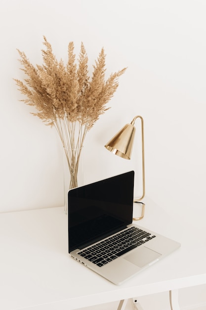 Computer portatile sul tavolo bianco con lampada dorata e erba di pampa