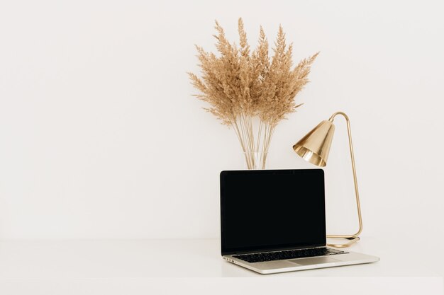 Photo laptop on white table with golden lamp and pampas grass