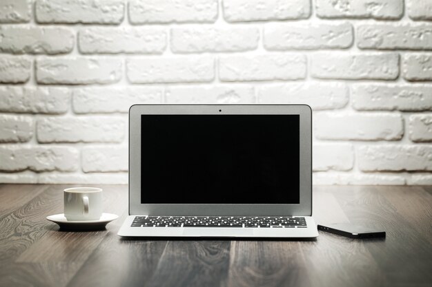 Laptop with blank black screen on a wooden table against white brick wall