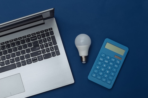 Ноутбук со светодиодной лампочкой, калькулятор на классическом синем фоне. Экономия энергии. Вид сверху
