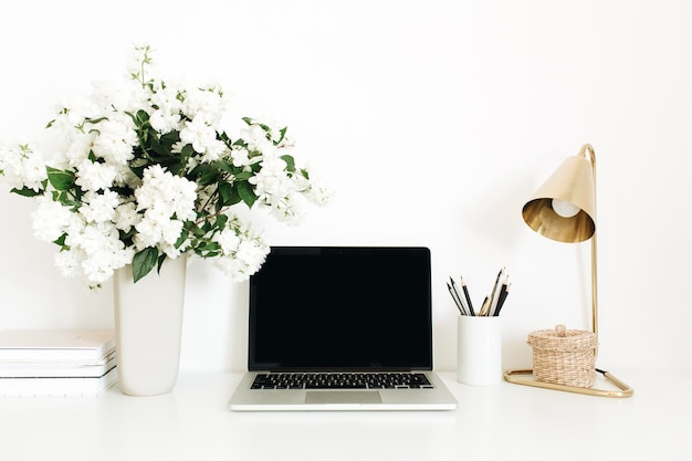 Laptop with mockup blank copy space screen on table. Office desk workspace with flowers bouquet, table lamp