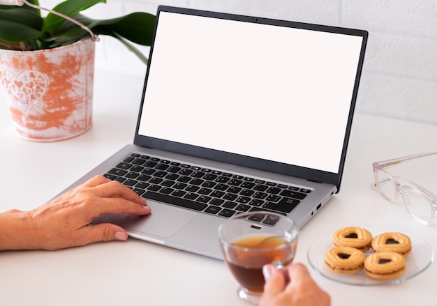 Laptop with white blank screen. female hand typing while
drinking a cup of tea. work place with pen and plant. technology,
internet, e-commerce, business