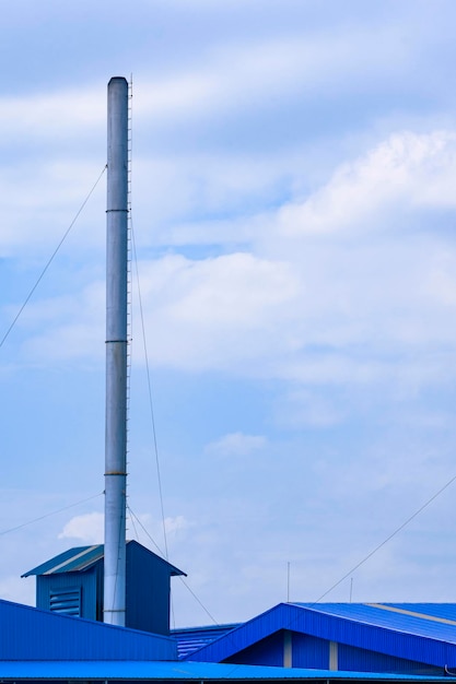 Large chimney on blue factory building roofs against white cloud and blue sky in vertical frame