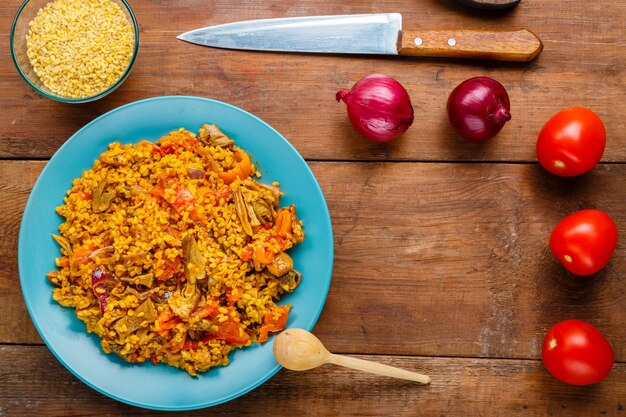 A large dish of bulgur with vegetables and mushrooms in spices on the table next to a knife