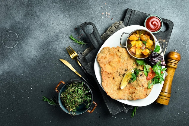 Большой жареный куриный шницель с овощами в тарелке на черном каменном фоне Фотография еды