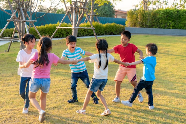 Large group of happy Asian smiling kindergarten kids friends holding hands playing and dancing play roundelay and stand in circle in the park on the green grass on sunny summer day.
