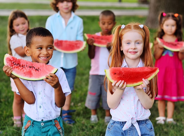 晴れた夏の日に、さまざまな民族の幸せな就学前の子供たちの大規模なグループが、熟したスイカのスライスを持って、公園の背景で笑っています。