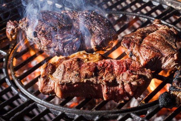 Large and juicy pieces of meat grilling on open fire Meat on metal grates covered by smoke Delicious and mouthwatering grilled food
