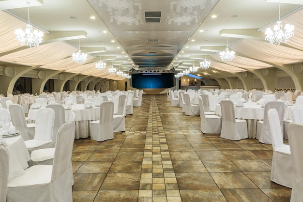 Большой освещенный банкетный зал, оформленный для светского мероприятия, со столами, сервированными посудой, и стульями в белых чехлах, роскошными подвесными люстрами с абажурами и сценой на заднем плане