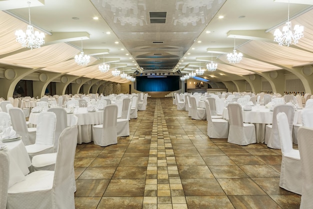 Photo large lighted banquet hall decorated for social event with tables served with dishware and chairs in white covers, luxurious hanging chandeliers with lampshades with stage in background