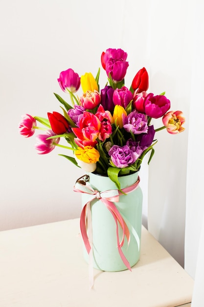 Large number of multicolor tulips in full bloom placed in light green metal vase with pink tape on neck, standing on white table on white background