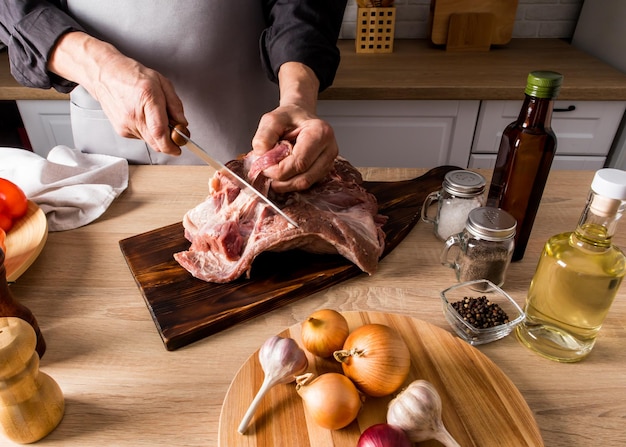A large piece of farm meat is cut by the cook on a kitchen board and a wooden countertop with ingredients for cooking