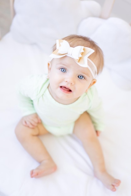 A large portrait of a cute little baby girl with big blue eyes looking into the camera with a bow on her head on a crib in a green cotton bodysuit