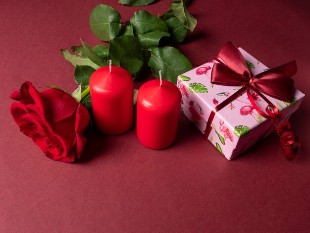 A large red rose that is next to two red large candles and a pink gift