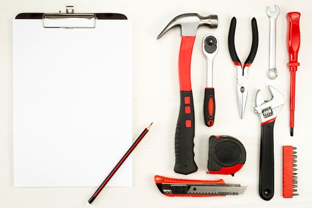 Large set of construction tools with a sheet of white paper with a pencil on a white wall, top view