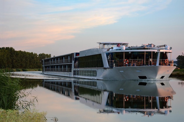 A large tourist passenger ship sails along the calm river between the banks