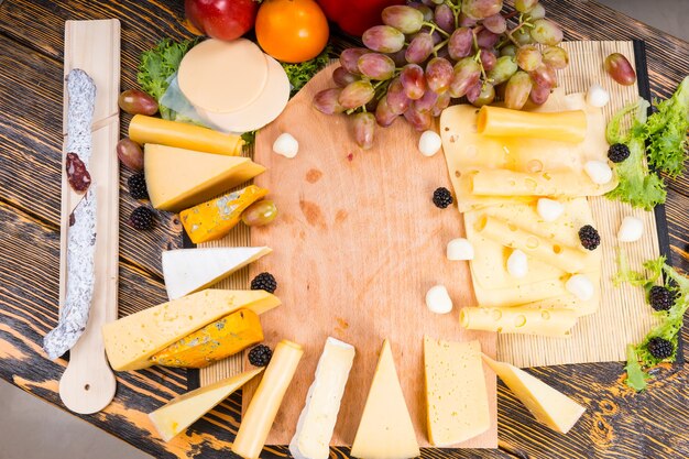 Large variety of cheeses displayed around a wooden cheeseboard with fresh grapes, tomato and spicy sausage on the side, overhead view with central copyspace