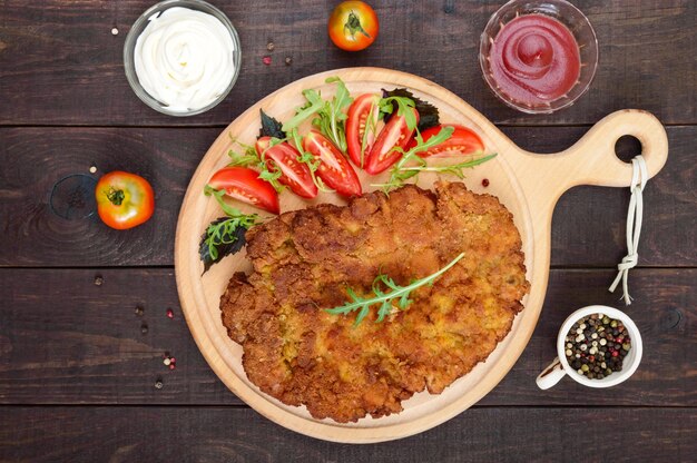 A large Viennese schnitzel  and tomato salad on cutting board on dark wooden table 