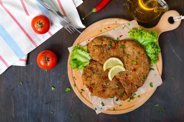 Large Viennese schnitzel on a wooden board with lemon on a dark background. Meat dish. Top view, flat lay.