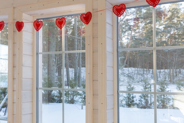 A large white bedroom window in wooden house decorated with romantic garland of red hearts on Valentine's Day. Bright interior of the room in a wooden house with a large window overlooking the winter