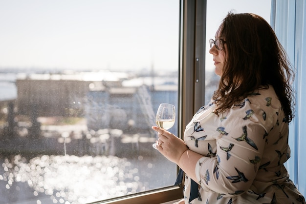 Large woman standing at the window with a glass of wine, the concept of loneliness