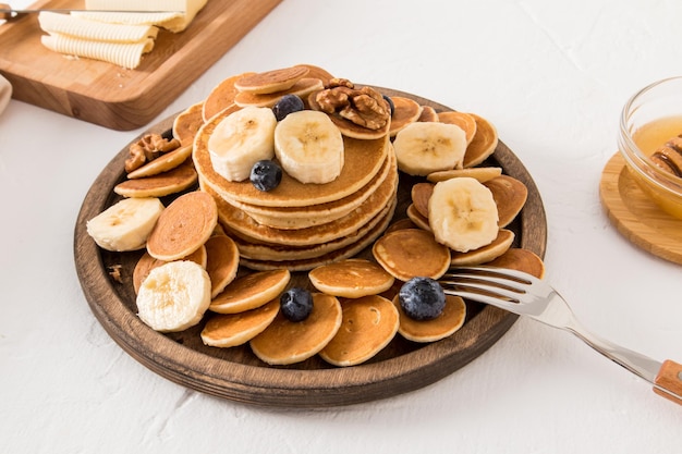 A large wooden plate with mini pancakes and a stack of thick pancakes in the middle banana slices fresh berries nuts on them white background