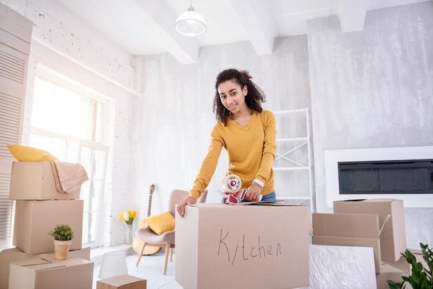 Foto ultimi preparativi. ragazza graziosa che sorride alla macchina fotografica e che chiude una scatola con piatti e utensili da cucina con nastro adesivo