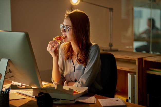 オフィスでの深夜の食事オフィスで遅くまで働いている間にピザを食べる魅力的な若い実業家のショット