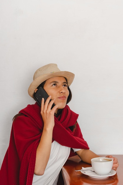 Latijnse boerin met hoed en een rode sjaal zittend die een kopje koffie drinkt terwijl ze luistert naar een bericht op haar mobiele telefoon op witte achtergrond