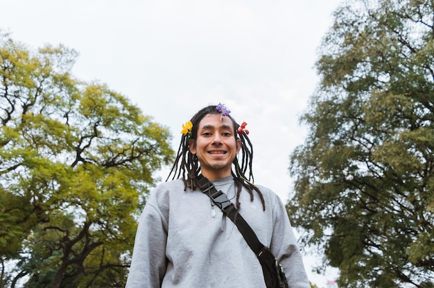 Latijnse jongeman met dreadlocks en bloemen op zijn haar die verlegen en glimlachend in het park staat