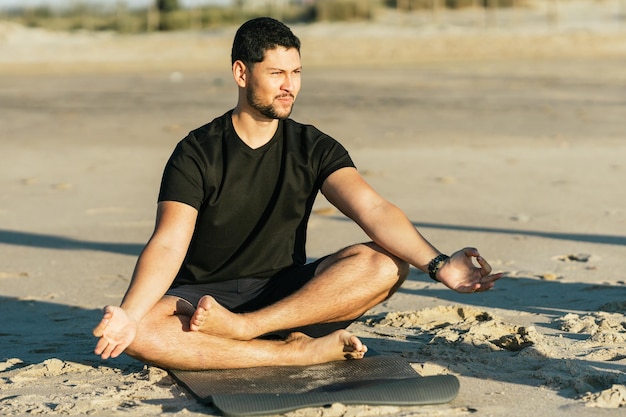 Latijnse man doet de lotuspositie van yoga met uitzicht op de zon zittend op een mat op het strand