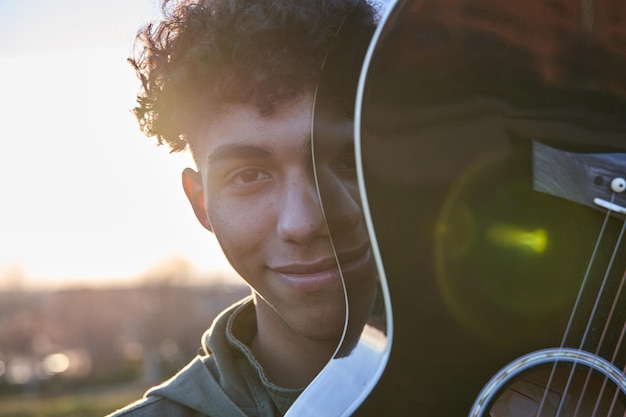 Latijnse tiener die zijn gezicht bedekt met gitaar Glimlachende jonge man met krullend bruin haar bij zonsondergang