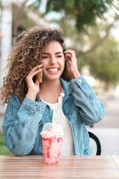 Latijnse vrouw die aan de telefoon praat terwijl ze een koud drankje drinkt in een coffeeshop. stedelijk begrip.