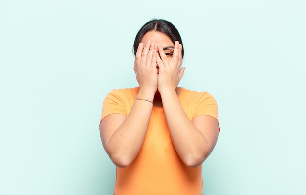 Latijnse vrouw die haar gezicht bedekt met handen, tussen de vingers gluurt met een verbaasde uitdrukking en opzij kijkt