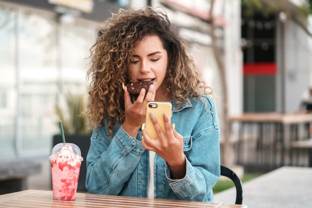 Latijnse vrouw die haar mobiele telefoon gebruikt terwijl ze een koekje eet en een koud drankje drinkt in een coffeeshop. stedelijk begrip.