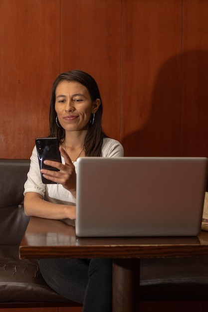 Latijnse vrouw die in een oude coffeeshop zit terwijl ze haar smartphone controleert en op haar laptop werkt