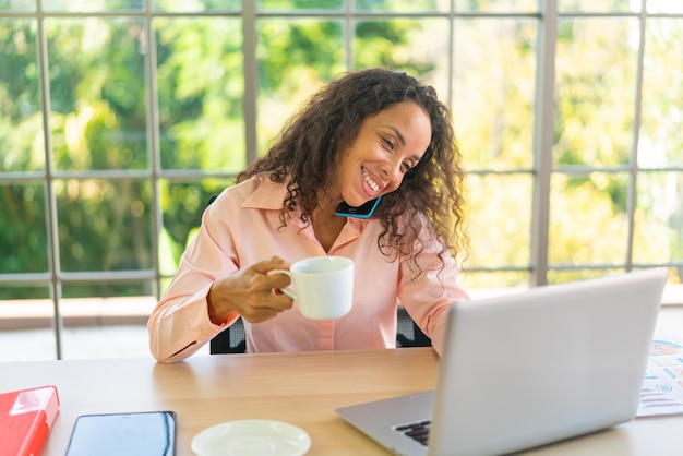 Latijnse vrouw die met koffiekop aan werkruimte werkt