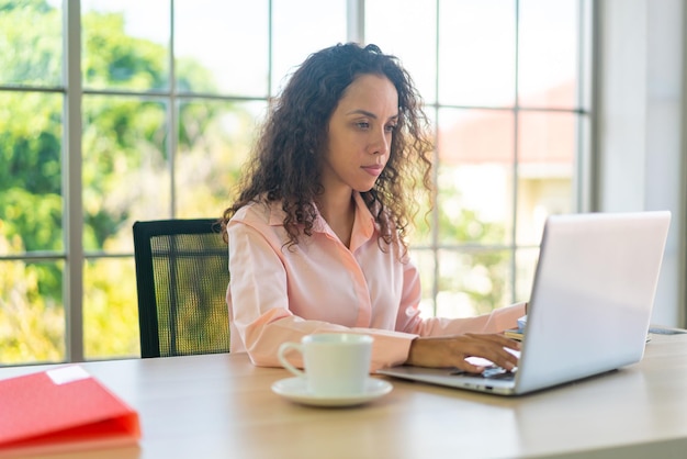 Latijnse vrouw die met laptop en papier aan werkruimte thuis werkt