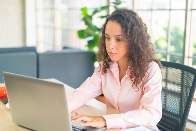 Latijnse vrouw die met laptop en papier aan werkruimte thuis werkt