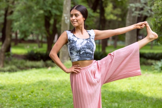 Latijnse vrouw die yoga doet, grijpt de voet in het park op groen gras met een roze outfit