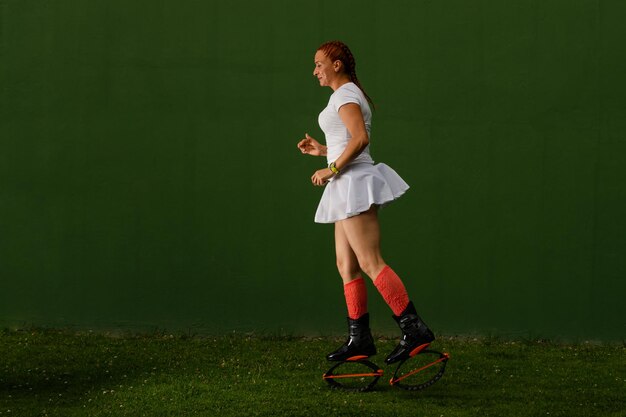 Latijnse vrouw die zijdelings staat in witte sportkleding en kangoo-springschoenen, één been naar achteren, op een donkergroene achtergrond