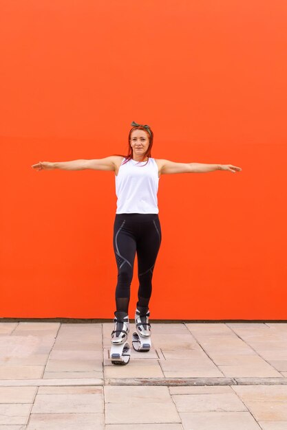 Latijnse vrouw gekleed in sportkleding en kangoo springt laarzen poseren met armen wijd open op een oranje achtergrond