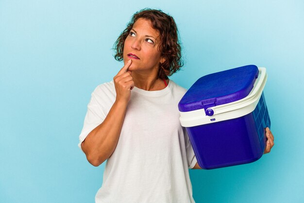 Latijnse vrouw van middelbare leeftijd geïsoleerd op blauwe achtergrond zijwaarts kijkend met twijfelachtige en sceptische uitdrukking.